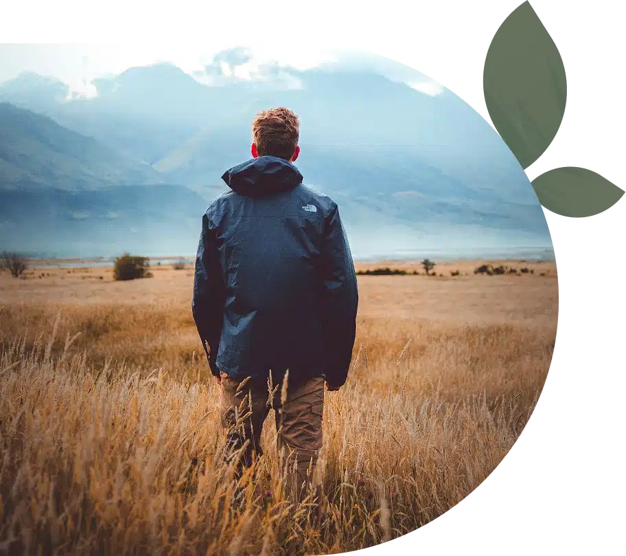 Person wearing a dark jacket standing in a golden field, facing distant mountains under a cloudy sky