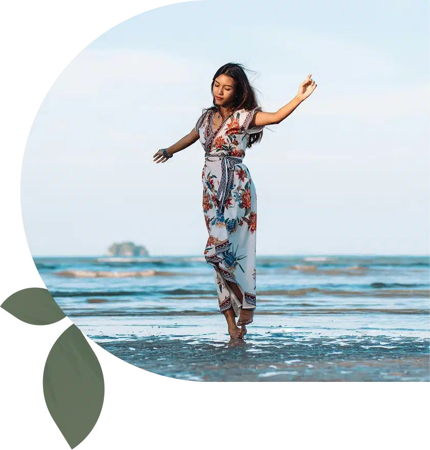 Woman in a floral dress walking barefoot on the beach, arms outstretched, with the sea and sky in the background